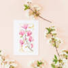 Carte illustrée d'une branche de magnolias roses en aquarelle, fond blanc, cadre doré, style floral délicat